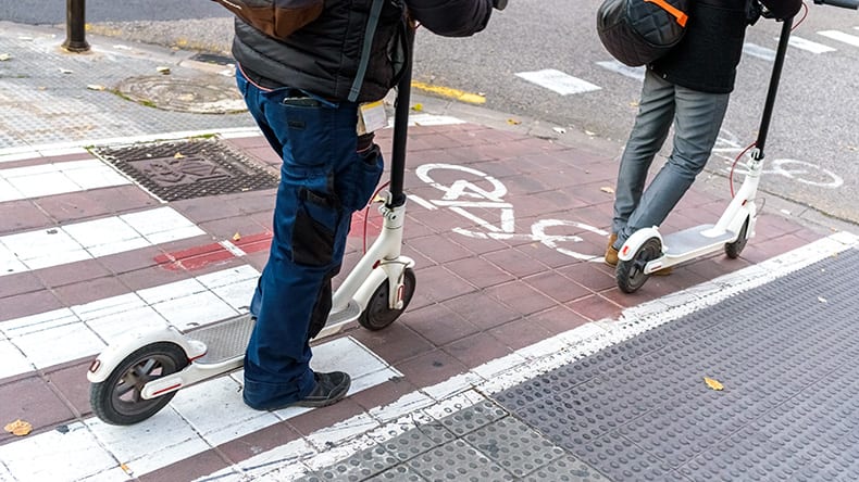 Two people ride electric scooters on a sidewalk near a bicycle lane. One person is wearing blue jeans, the other grey pants. The pavement is patterned with red and beige tiles and a marked bike symbol.