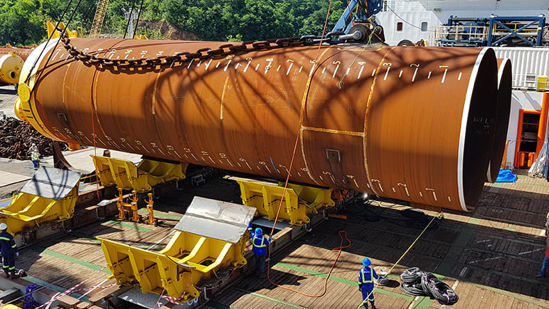 Workers in safety gear stand near a large, rust-colored cylindrical pipe being lifted by a crane at an industrial site. Trees and equipment containers are visible in the background.