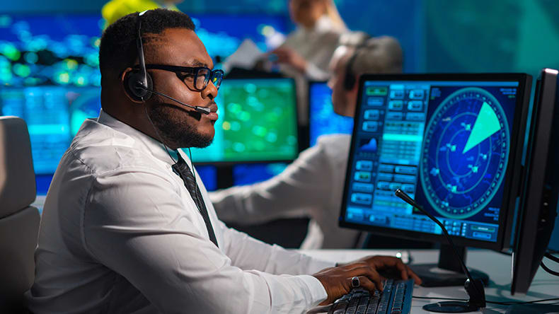 A man in a white shirt and headset monitors data on multiple screens in a control room, focusing on radar displays. Another person in the background works at a similar station.