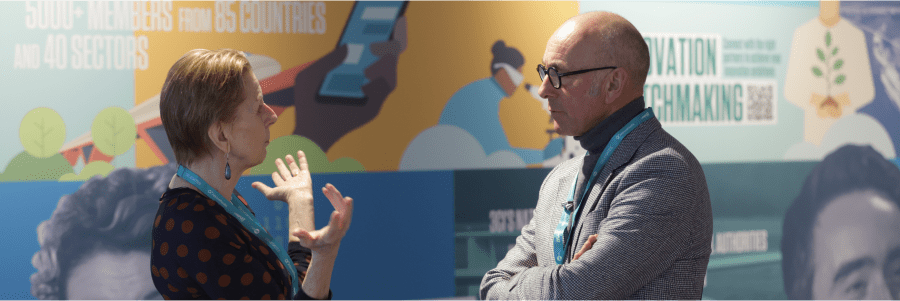 Two people engaged in conversation at an event, standing in front of a colorful display with text and images. Both are wearing event lanyards, and their expressions suggest an animated discussion.