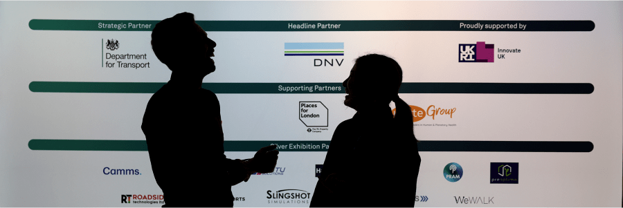 Silhouettes of two people in conversation with a backdrop displaying various company logos and organizations including the Department for Transport, DNV, and Innovate UK.