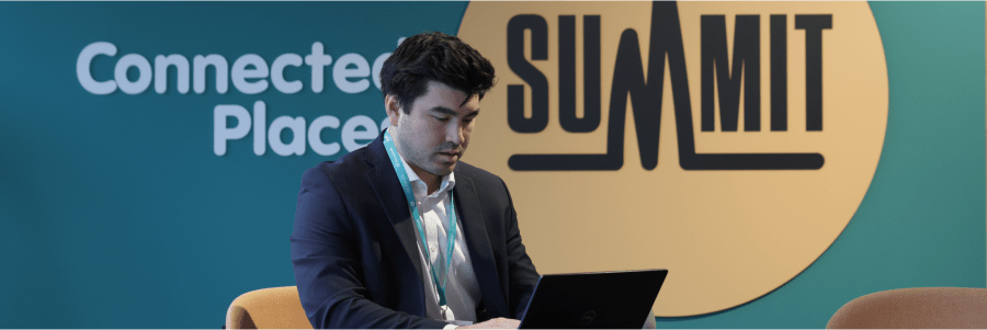 A man in a navy suit sits in front of a blue backdrop with "Connected Places Summit" text, working on a laptop.