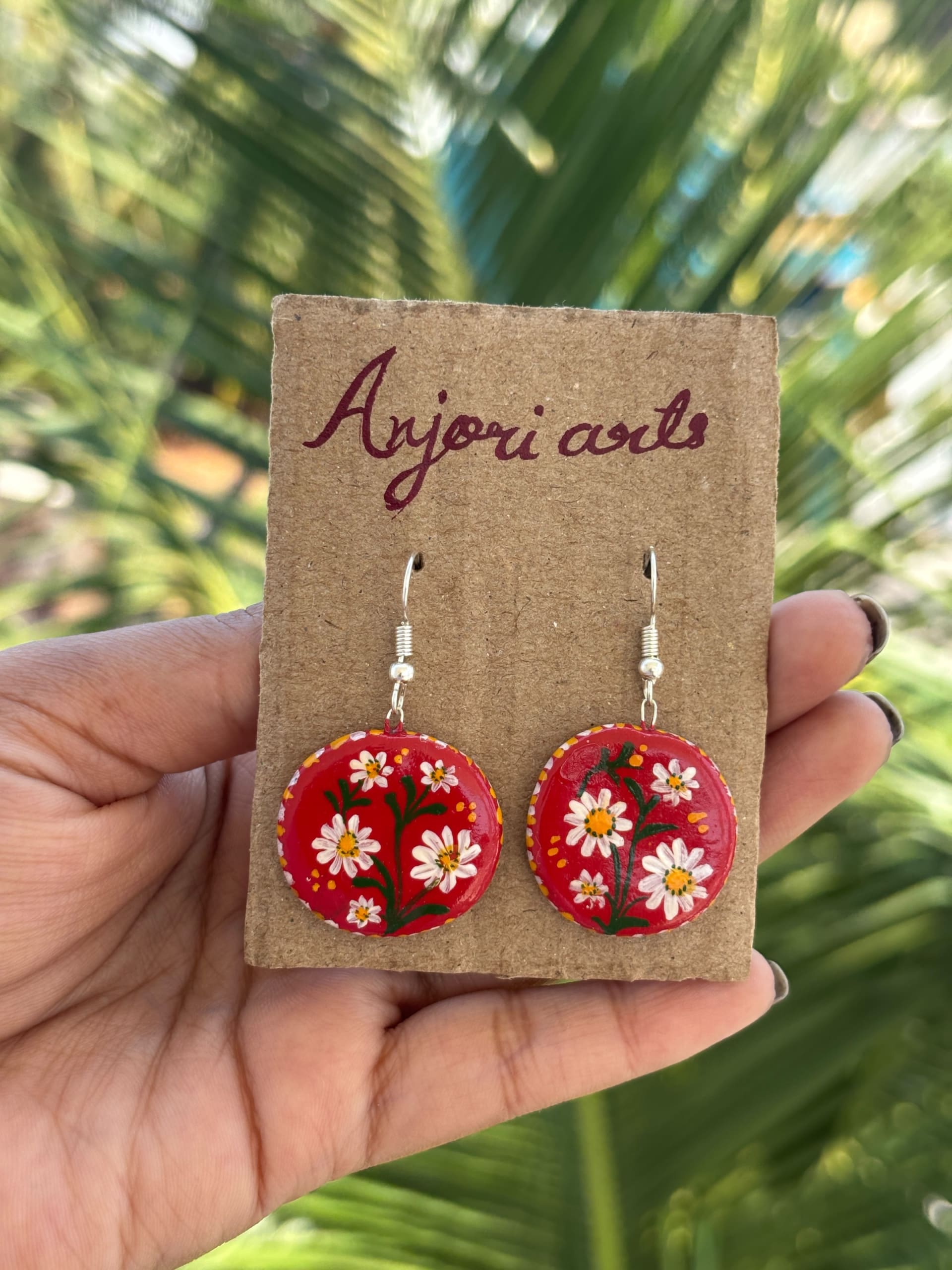 A pair of round wooden earrings painted with white daisies on a red background, held by a hand on a brown cardboard label.