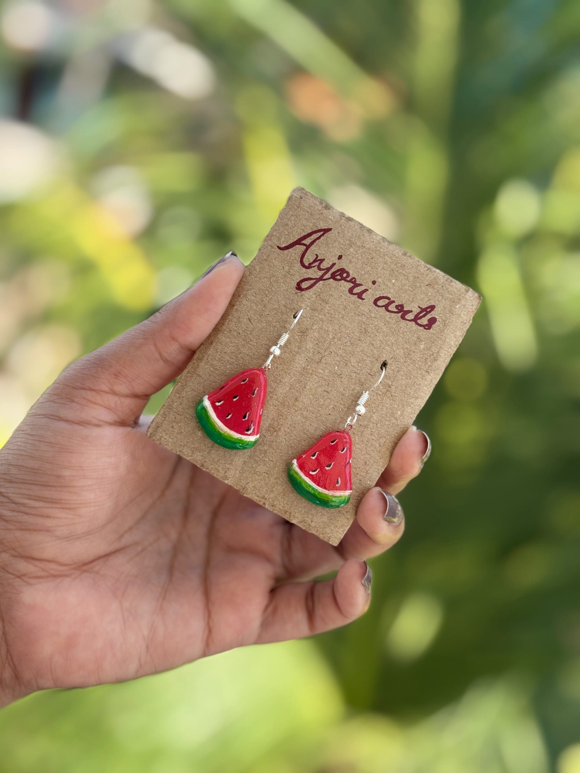 Hand-painted watermelon slice earrings in red, white, and green on a cardboard display card, held by a hand.