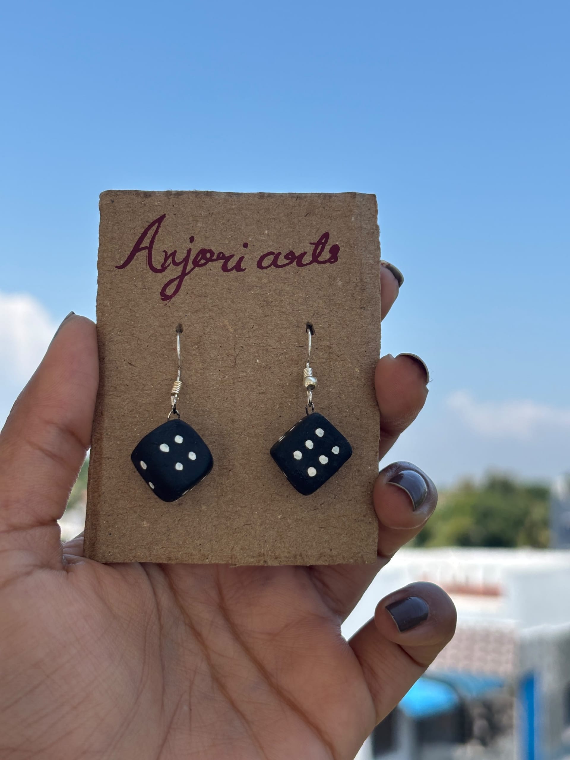 A hand holding a pair of black dice-shaped earrings with white dots on a brown cardboard display card against a blue sky.