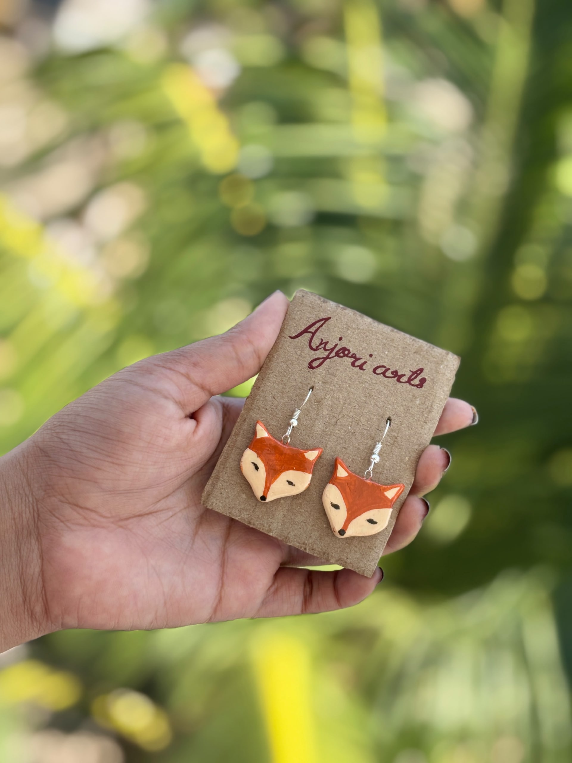 A hand holding a pair of hand-painted fox-shaped clay earrings on a brown display card with Anjori Arts branding.