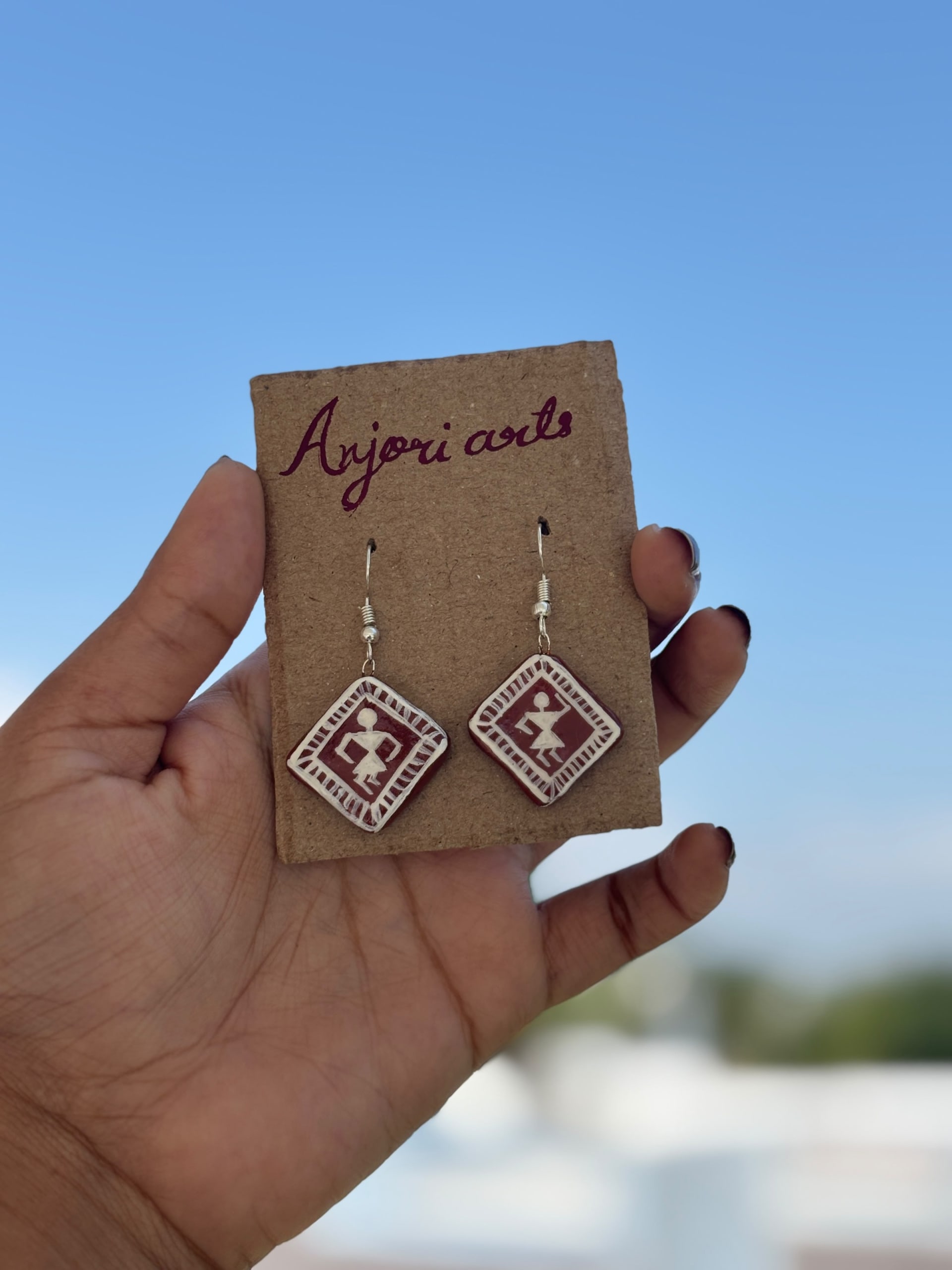 A hand holding two diamond-shaped terracotta earrings featuring white hand-painted Warli tribal figures on a clear blue sky.
