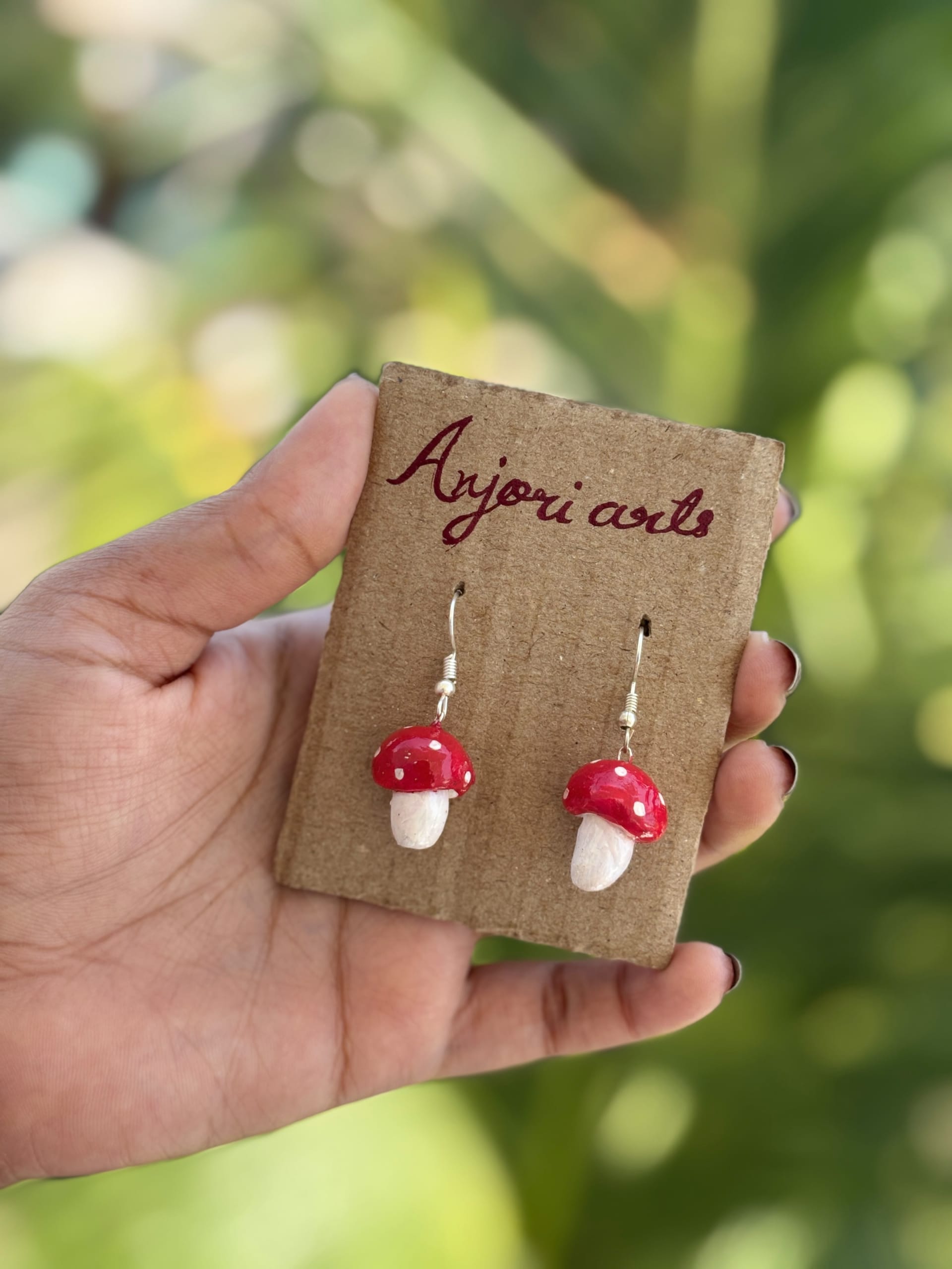 A pair of hand-sculpted red and white toadstool clay earrings on a branded Anjori Arts cardboard display card.