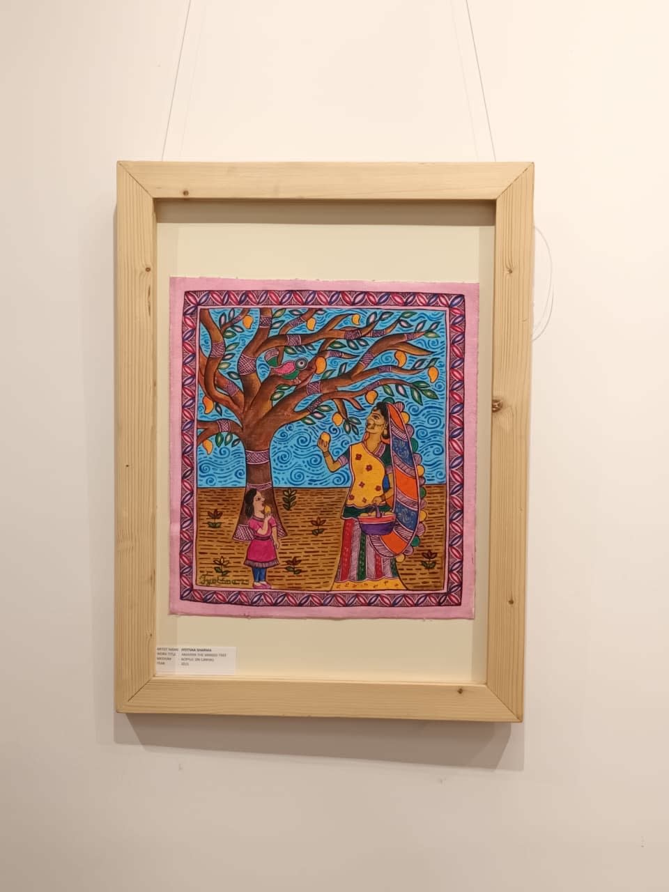A colourful folk-style painting in a wooden frame shows a woman and child gathering yellow mangoes beneath a tree against a blue sky.