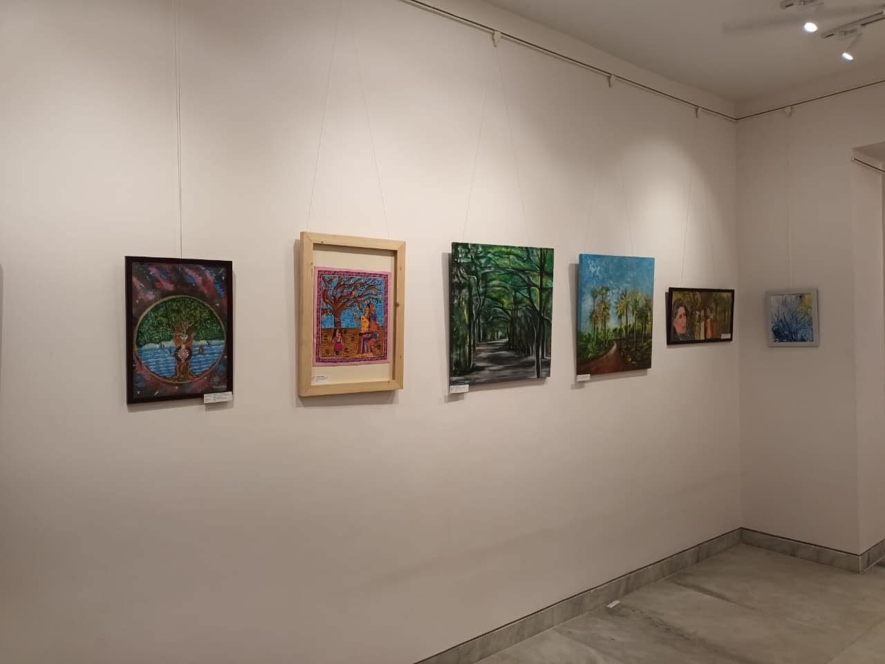 Several framed artworks hanging on a white wall in a gallery, featuring landscape paintings, a circular tree motif, and figures.