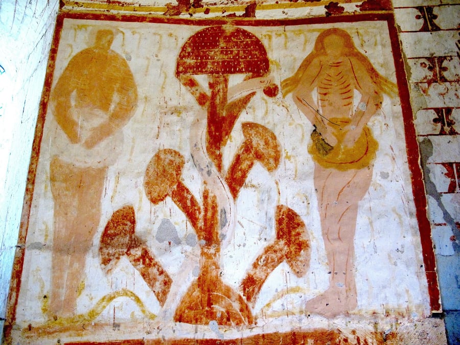 Faded medieval wall painting in red and ochre. Adam stands on the left and a thin Eve with painted ribs on the right. Between them is a tree shaped like a giant dotted mushroom cap with four smaller caps below, with a pale serpent wrapped around the trunk holding a small red fruit toward Eve.