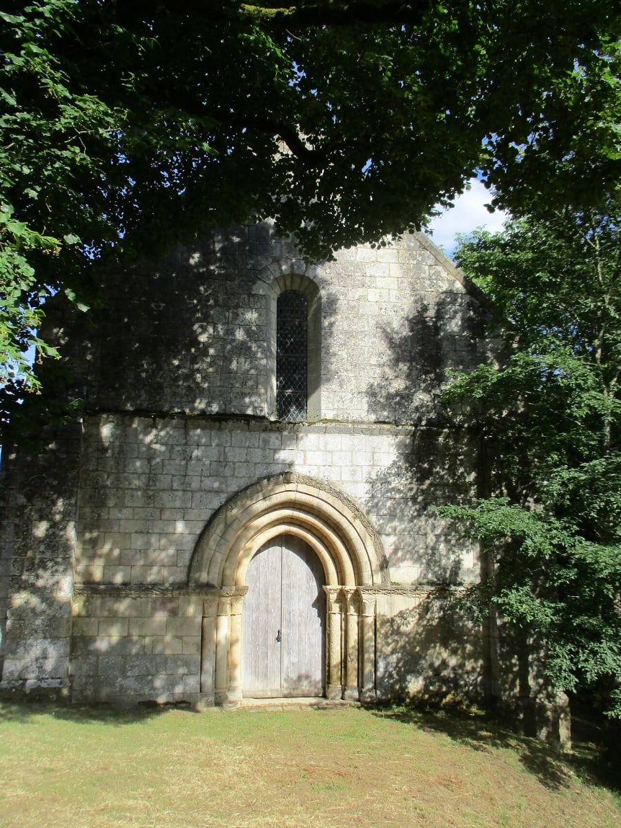 The stone west front of a small Romanesque chapel framed by trees, with a round-arched carved doorway and a single narrow window above.