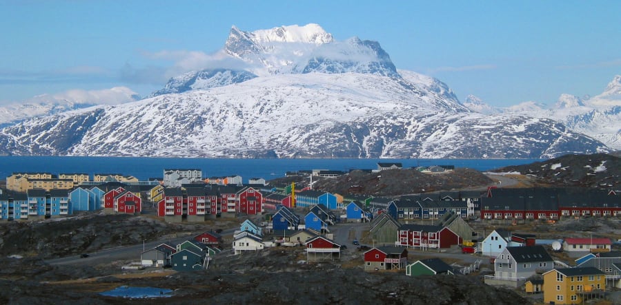 Colourful wooden houses and apartment blocks of Nuuk on a rocky hillside, with a fjord and a snow-covered mountain behind them under a blue sky.