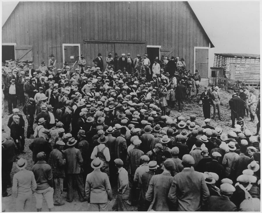 Black-and-white photo of hundreds of men in caps and hats packed in front of a large wooden barn, with men in uniform, some holding clubs, lined up along the barn wall.