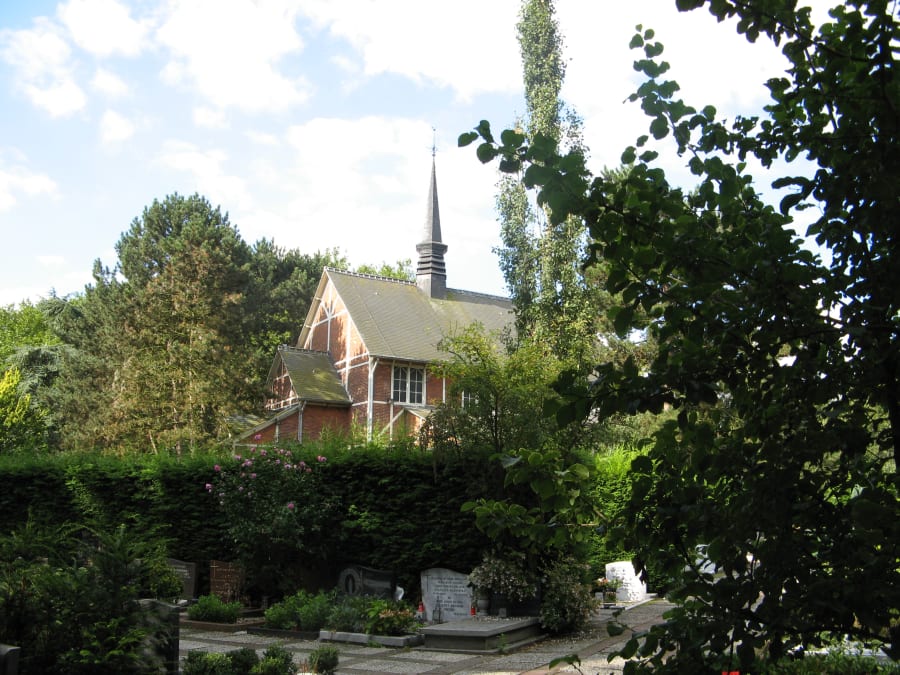 Graves and flowering shrubs in the foreground of a leafy cemetery, with a red-brick chapel and a slender spire behind a tall hedge.