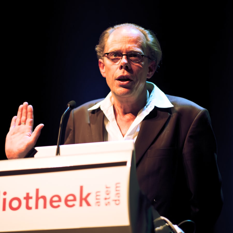 A man with glasses and thinning hair, in a dark jacket and open-necked white shirt, speaks at a lectern with one hand raised.