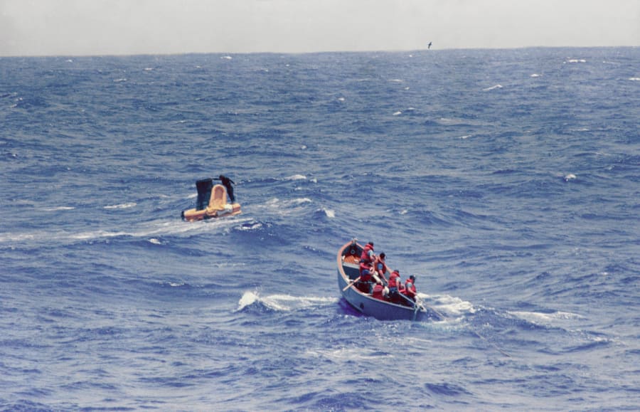 A small capsule wrapped in an orange flotation collar bobs in choppy blue sea, with a swimmer standing on it, while a rowing boat full of sailors in orange life jackets approaches.