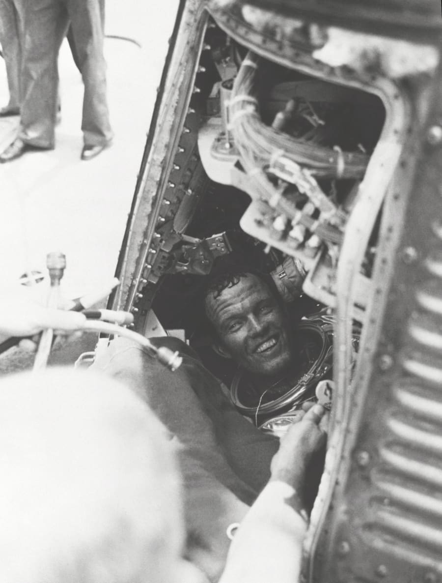 A smiling man with tousled hair, in a silver pressure suit with a red collar, sits inside the open hatch of a capsule surrounded by equipment.