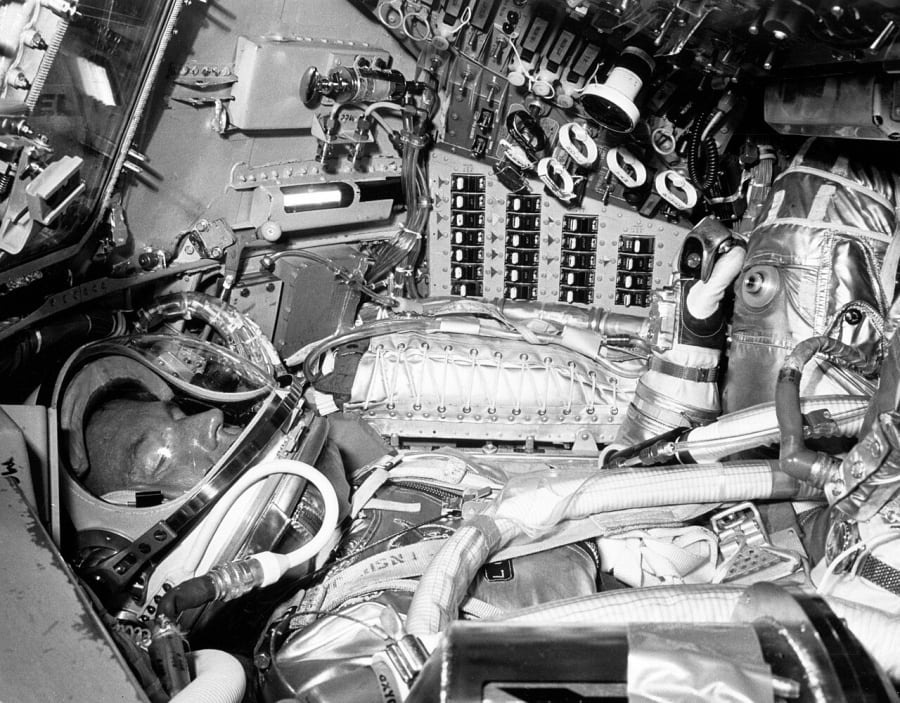Black-and-white photo of an astronaut in a helmet and silver pressure suit lying in a cramped capsule, surrounded by rows of switches, dials, hoses and a window frame at upper left.