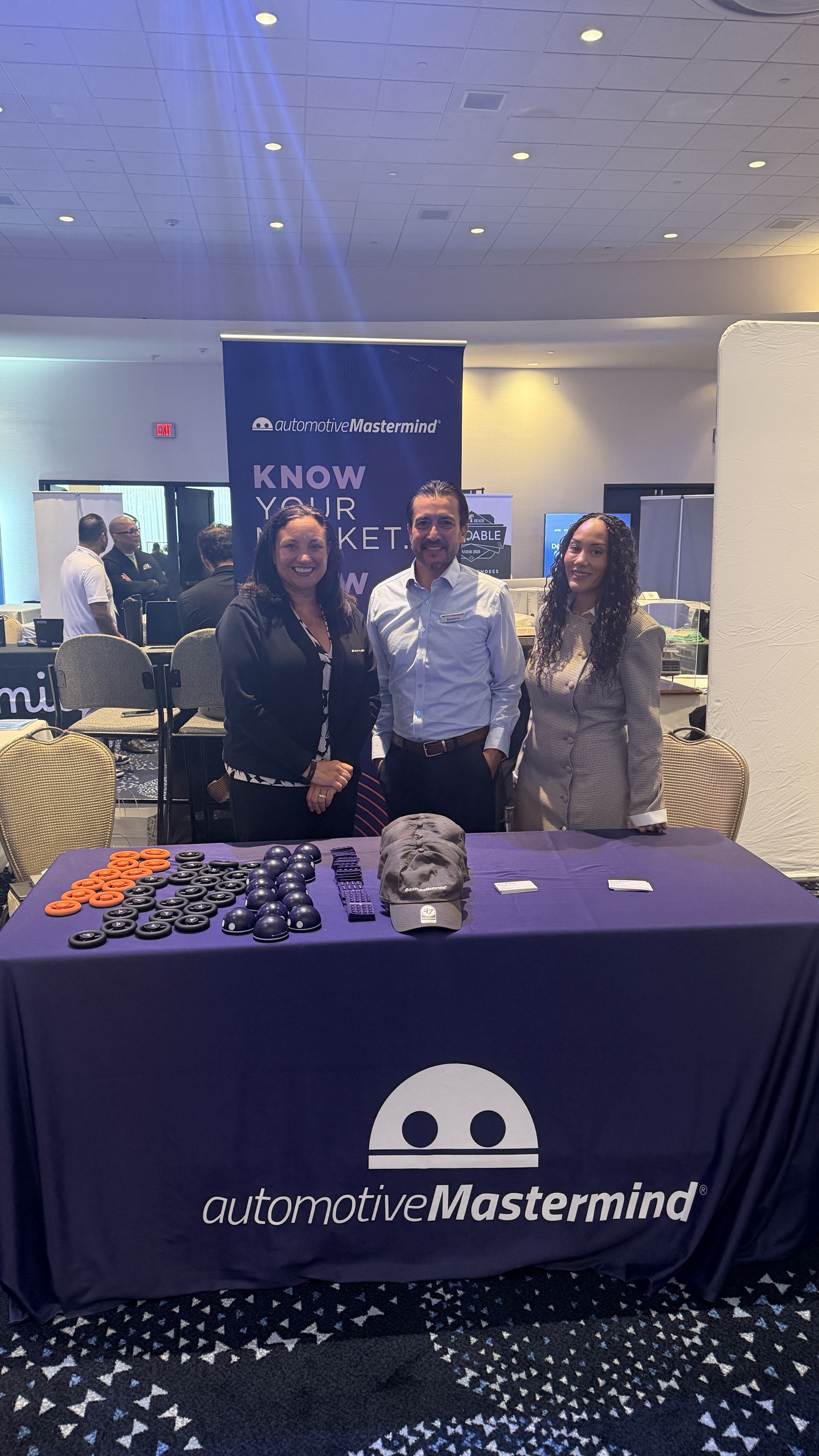 Three employees standing in front of a branded automotiveMastermind table holding giveaway promotional items. They are standing in front of a tall banner in a banquet hall.