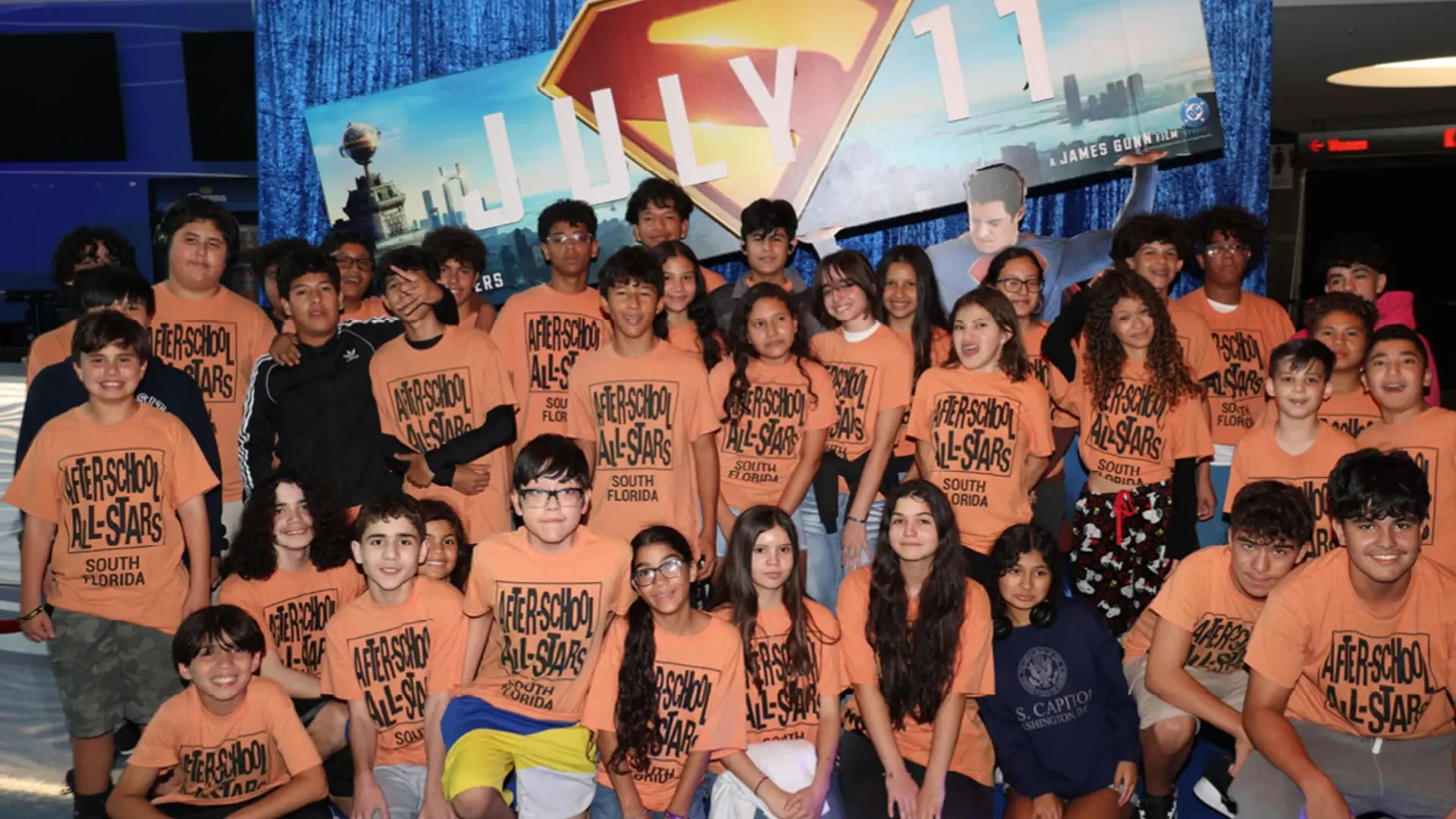 A large group of students wearing orange shirts stand in front of a superman symbol