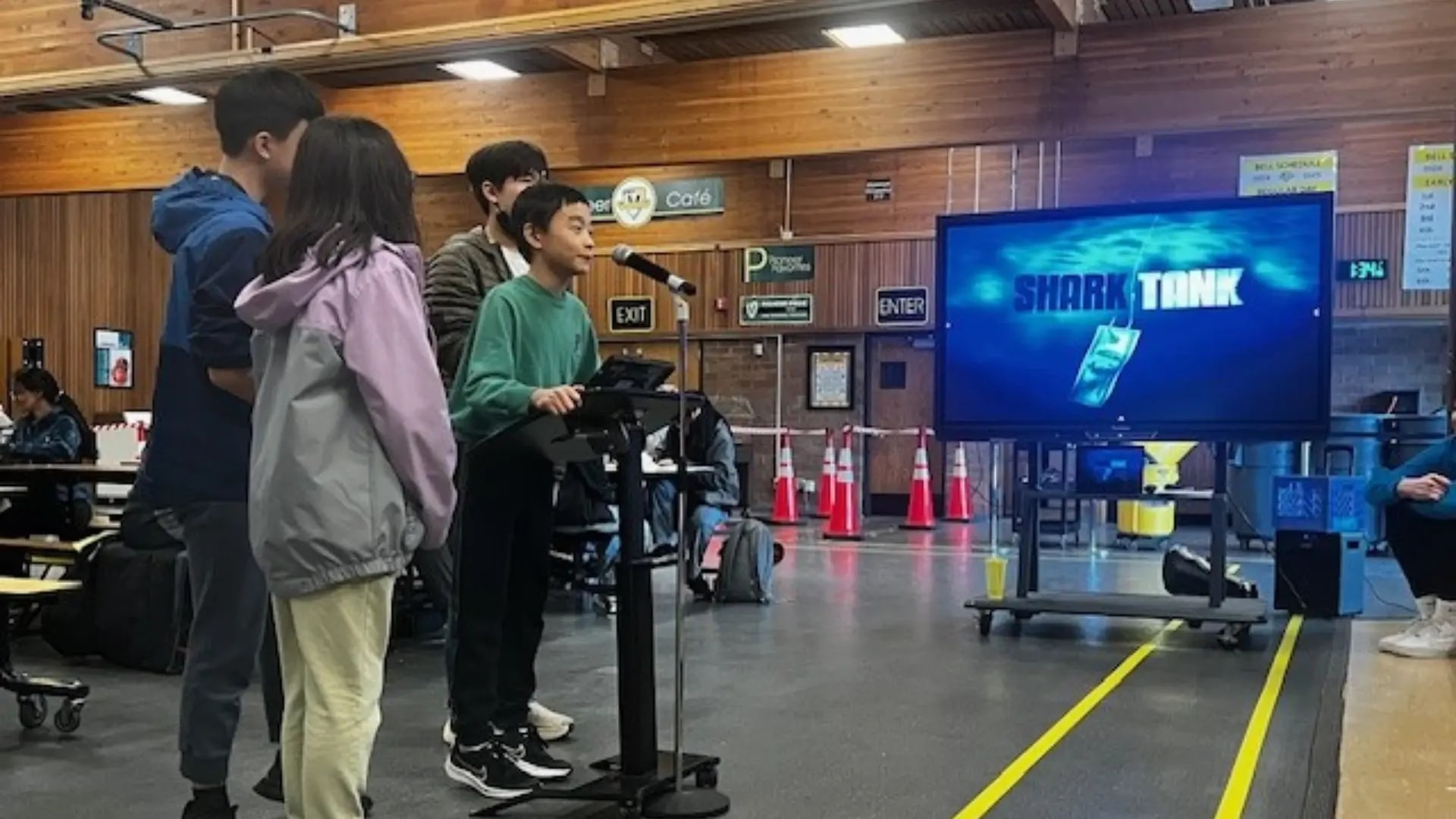 A young boy in a green shirt stands in front of a microphone. Three other students stand around him