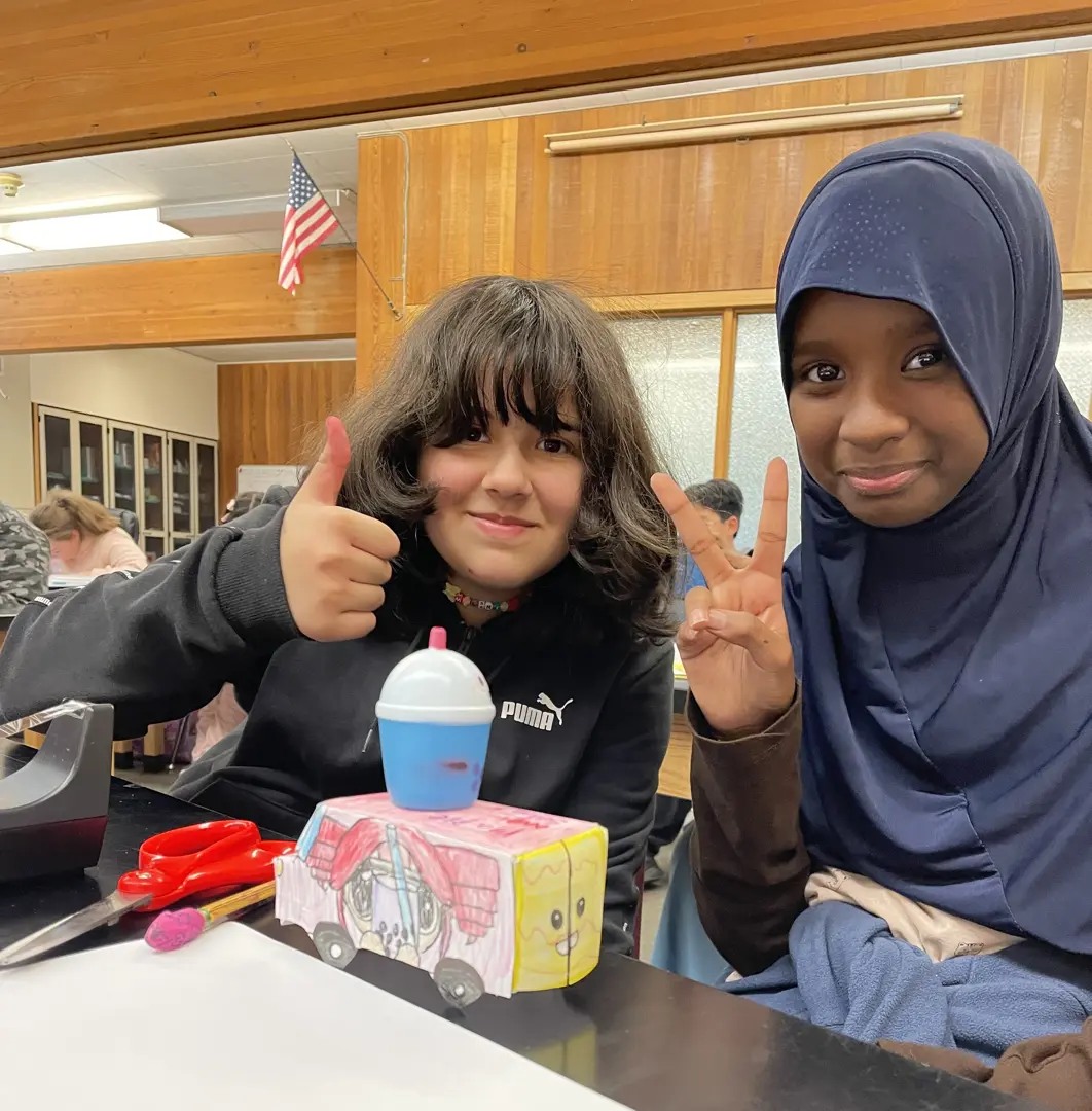 Two students, one wearing a black sweater and giving a thumbs up, one wearing a blue hijab and holding up a peace sign.