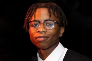 A student wearing a black shirt with a white collar and glasses against a black background
