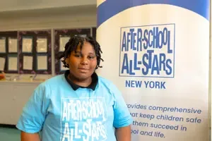 A student wearing a blue After School All Stars shirt stands against an ASAS backdrop