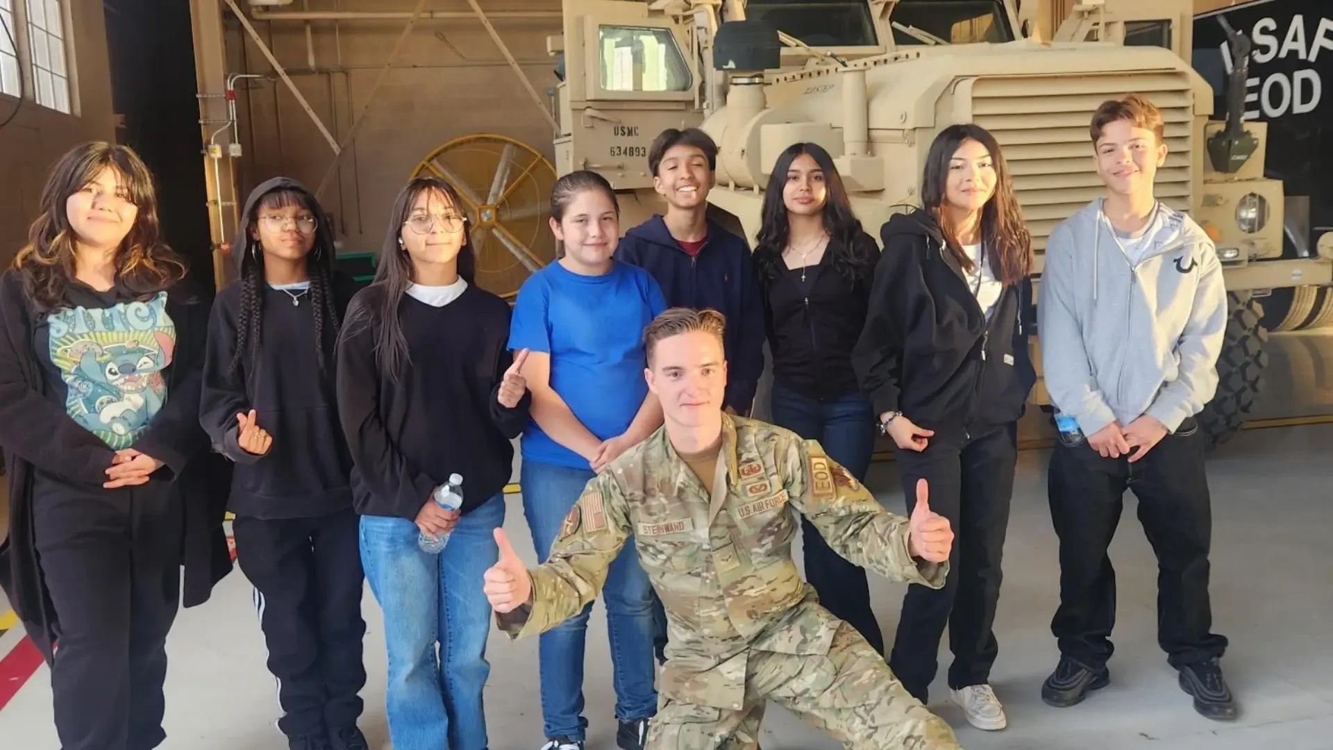 A man in Air Force uniform kneels and gives two thumbs up in front of a group of students