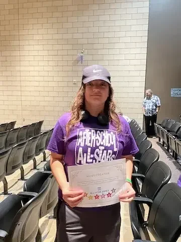 A girl in a purple shirt holds a piece of paper and stands in front of a row of chairs
