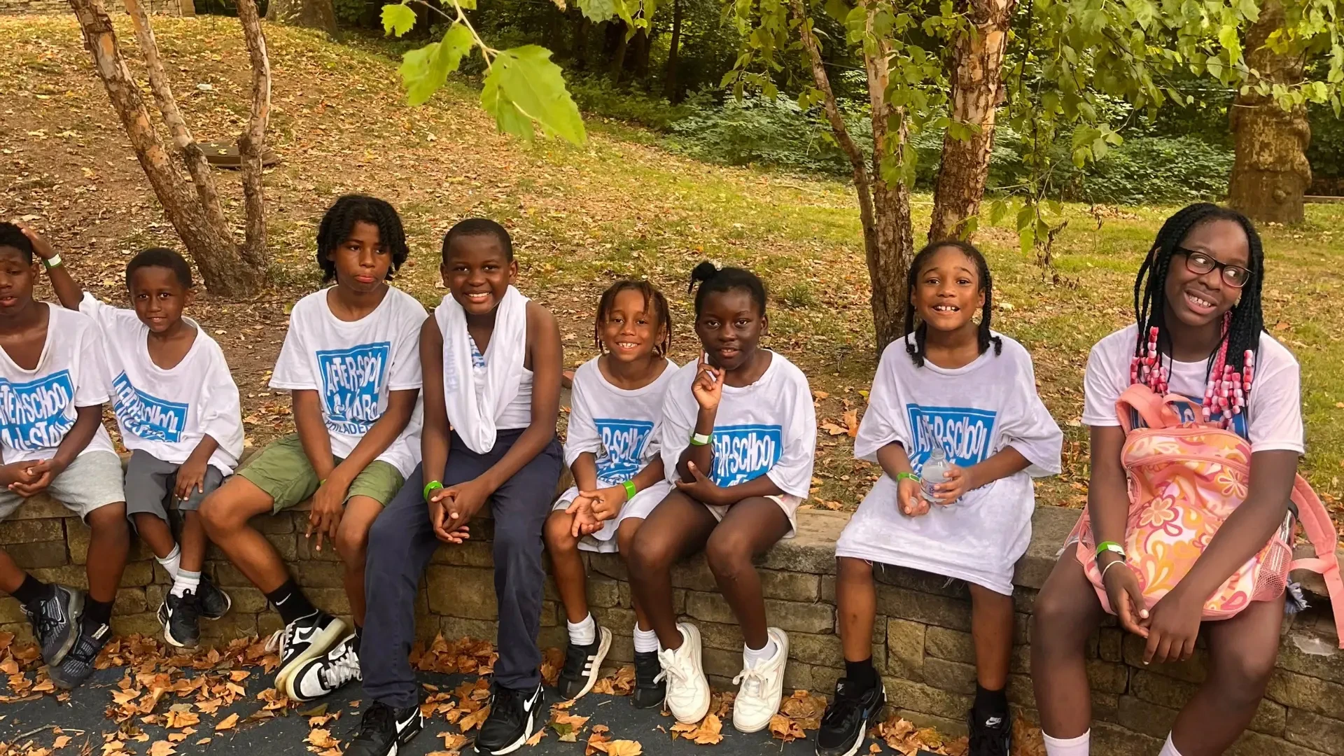 A group of students in matching white After-School All-Stars shirts sit on a rock wall
