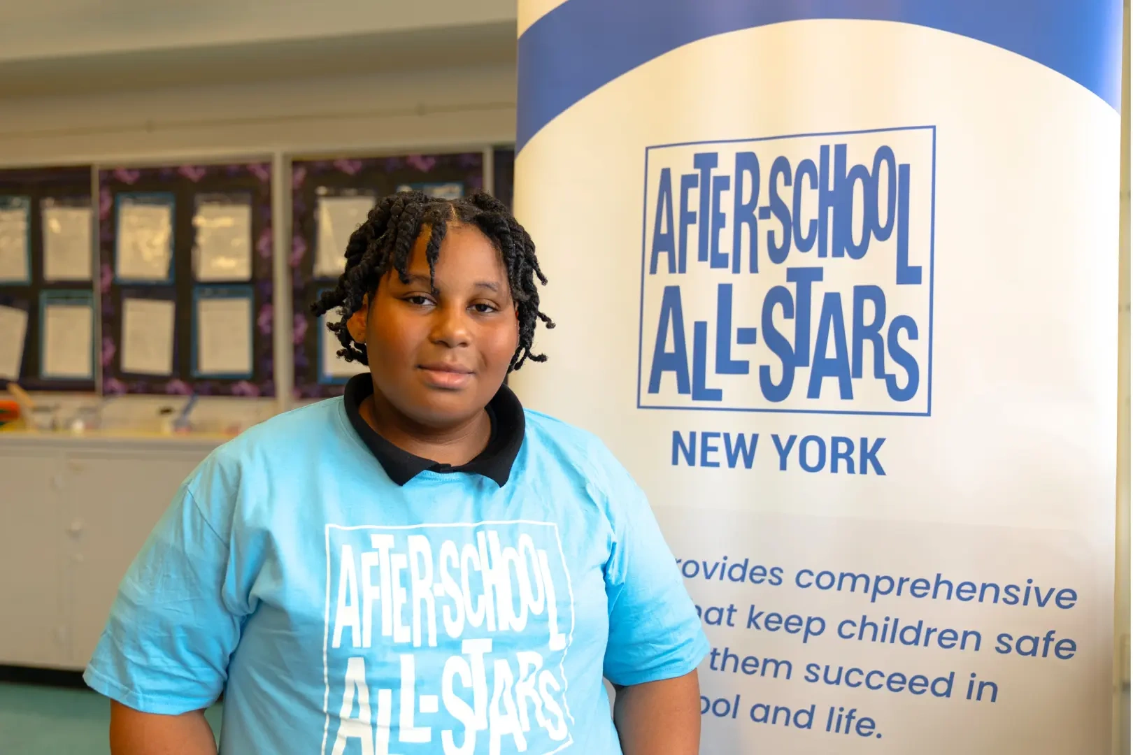 A young boy wearing a blue shirt stands in front of the After-School All-Stars logo