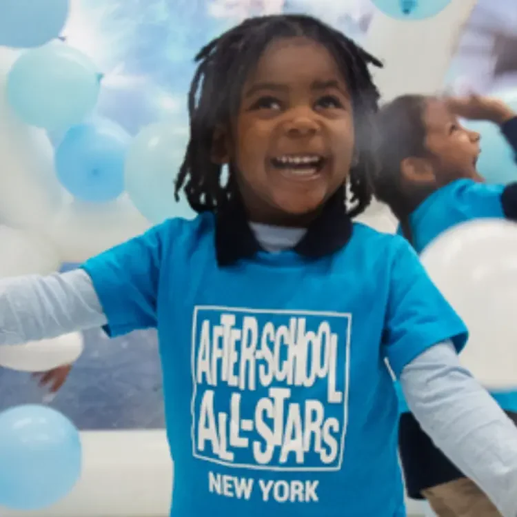 A young girl with a blue shirt smiles in front of a large amount of balloons