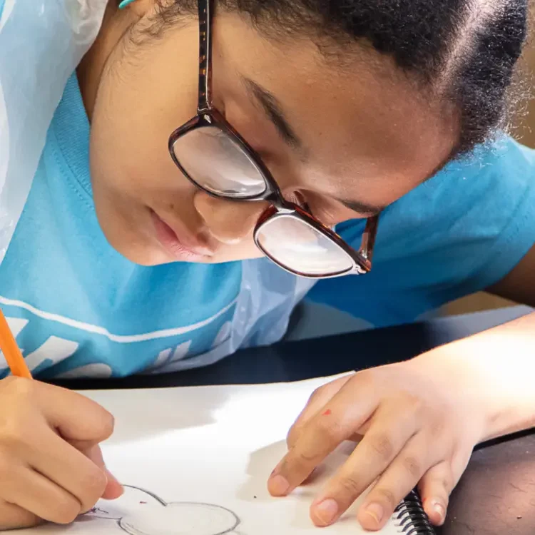 A student wearing glasses and a blue shirt writes on a piece of paper with a pencil