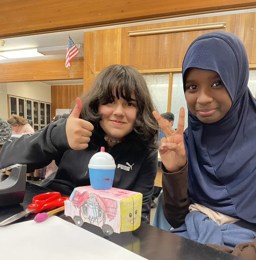 Two students, one wearing a black sweater and giving a thumbs up, one wearing a blue hijab and holding up a peace sign.