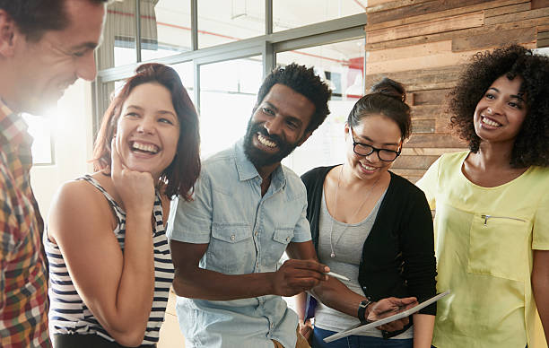 A diverse group of five people are gathered in a casual setting, smiling and engaged in conversation. One person holds a tablet, and they appear to be discussing something on the screen. The background shows a bright, modern interior.