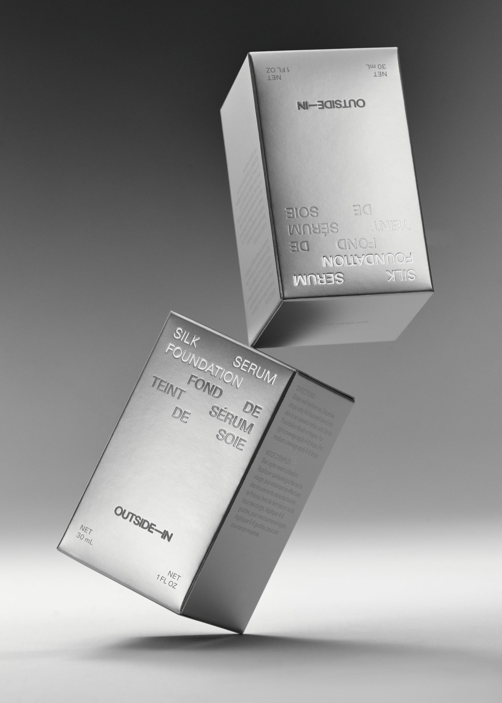 Two Outside-In Silk Foundation Serum metallic silver boxes suspended in the air at angles against a soft grey gradient background