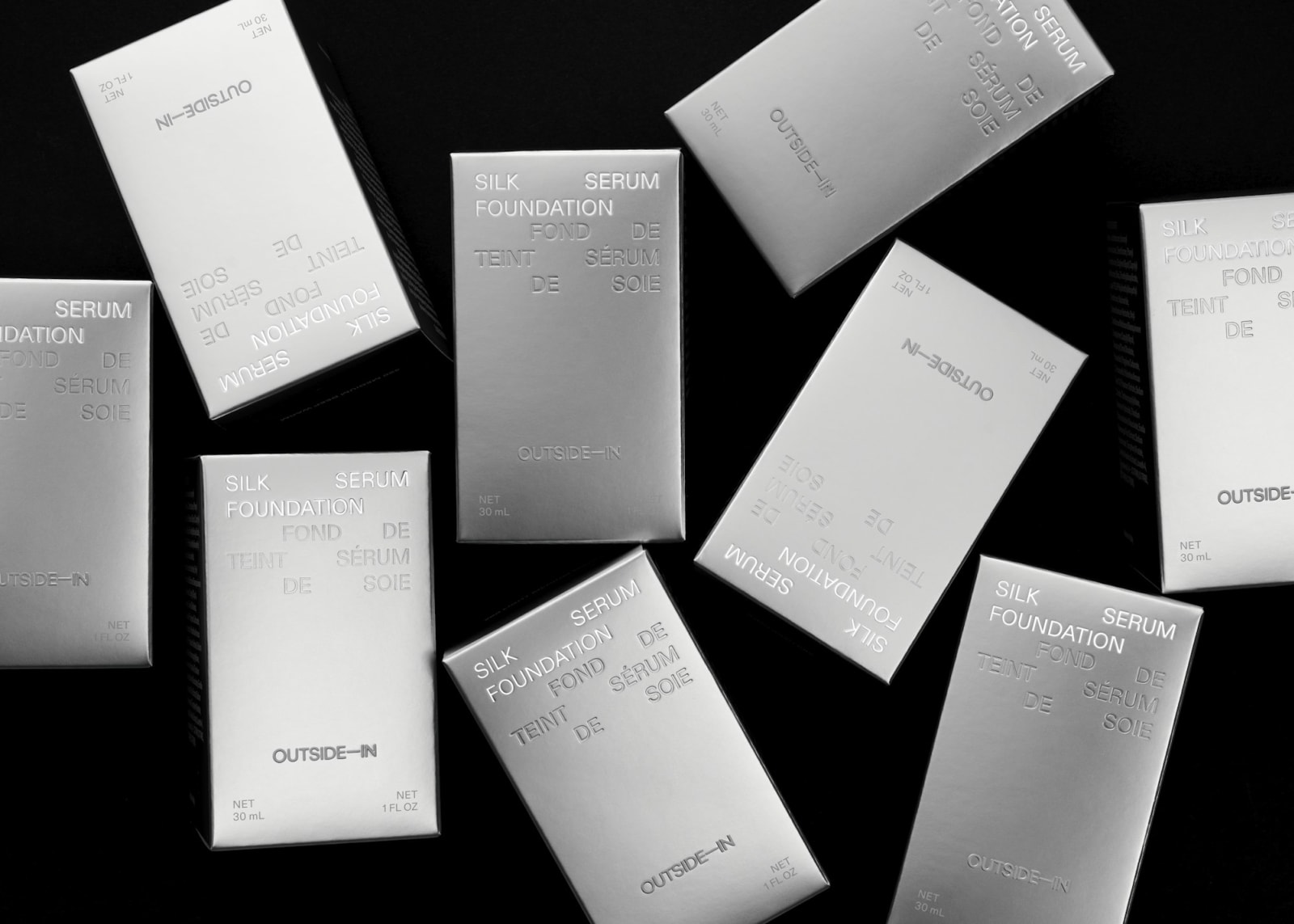 Multiple Outside-In Silk Foundation Serum metallic silver boxes scattered on black background in bird's-eye view