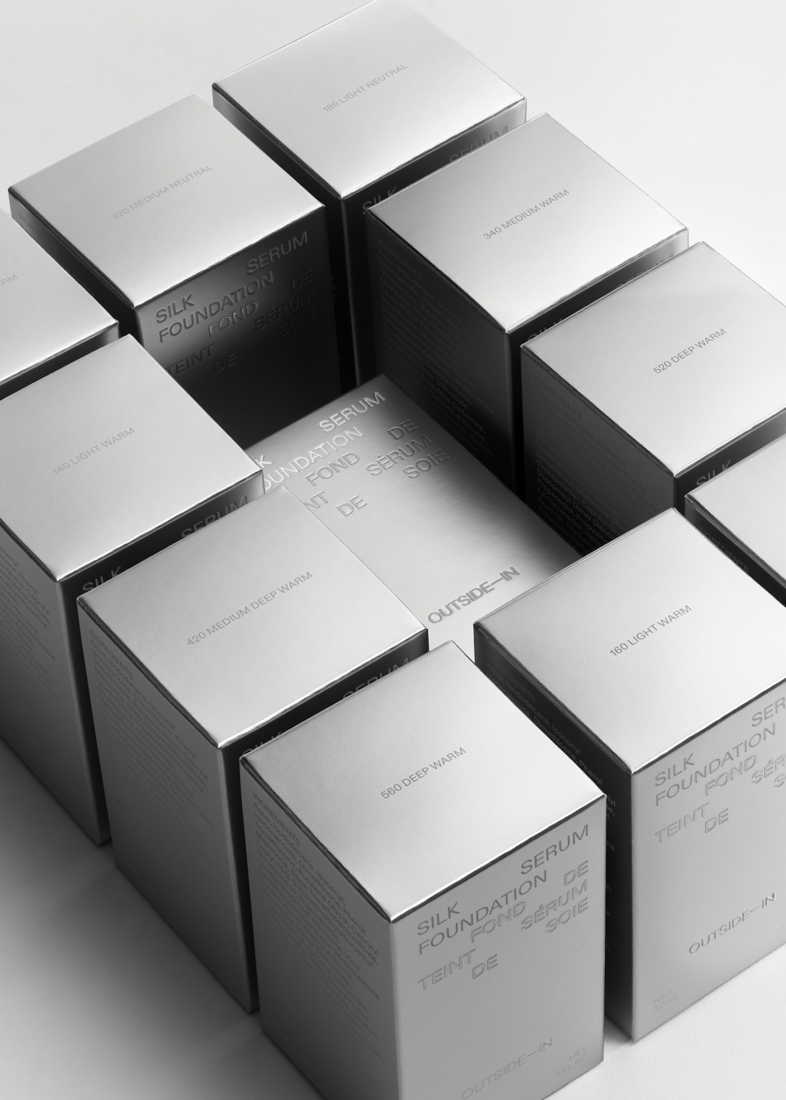 Overhead view of nine Outside-In Silk Foundation Serum metallic silver boxes scattered on white surface showing the full shade range