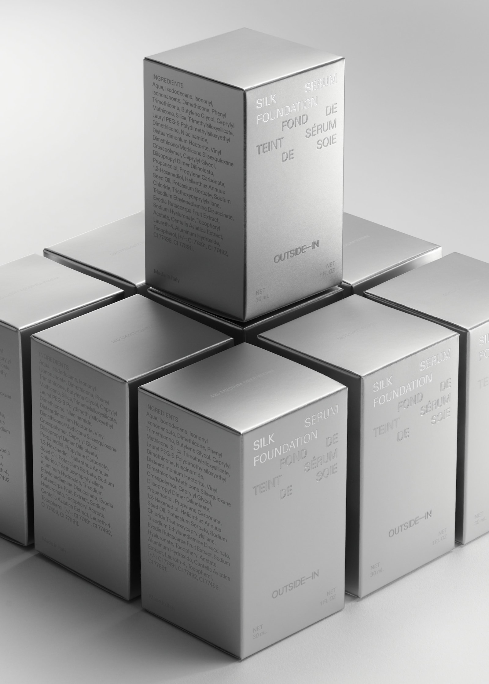 Outside-In Silk Foundation Serum metallic silver boxes arranged in a three-by-three grid with one box elevated at the top on grey background