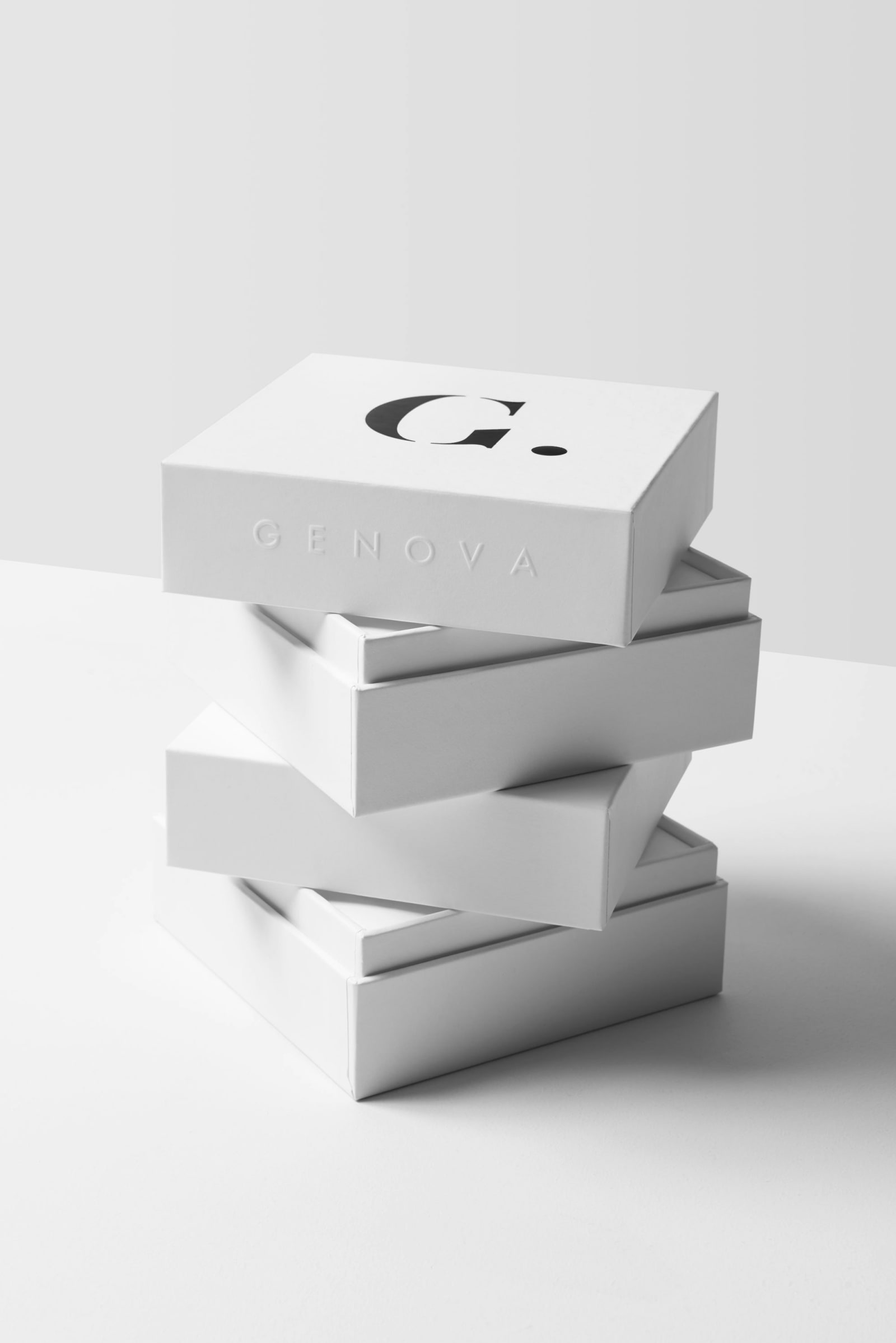 Four white rigid boxes stacked at offset angles with large black serif G. on top face and GENOVA blind-embossed on side against white background