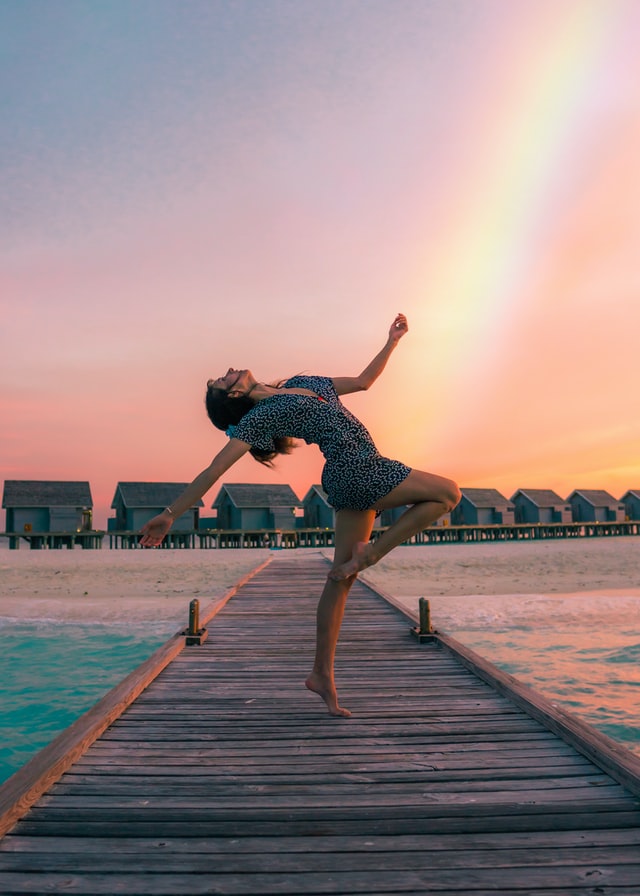 woman dancing on a dock with a rainbow filled sky behind her