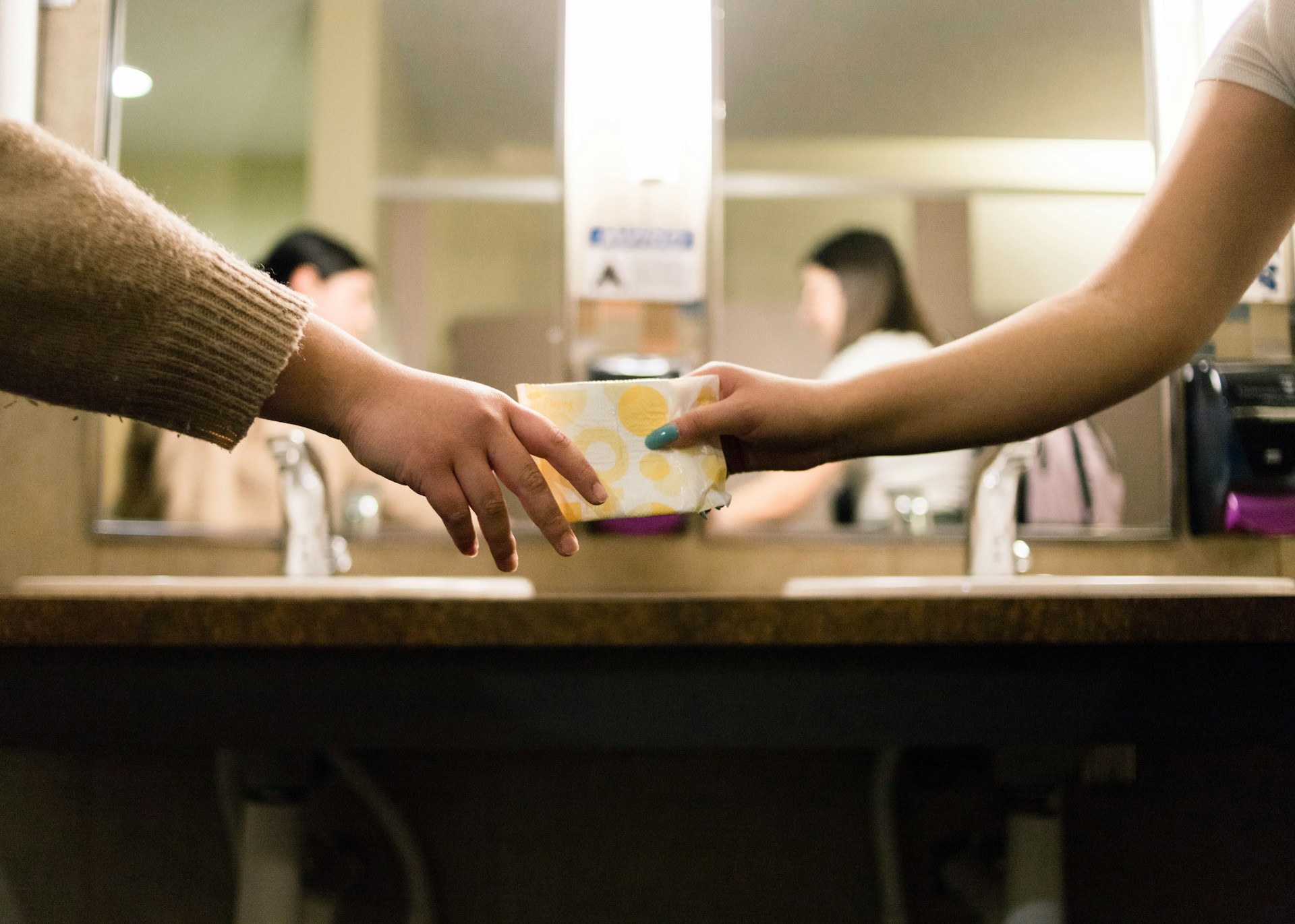 woman handing another woman a menstrual pad in a public bathroom