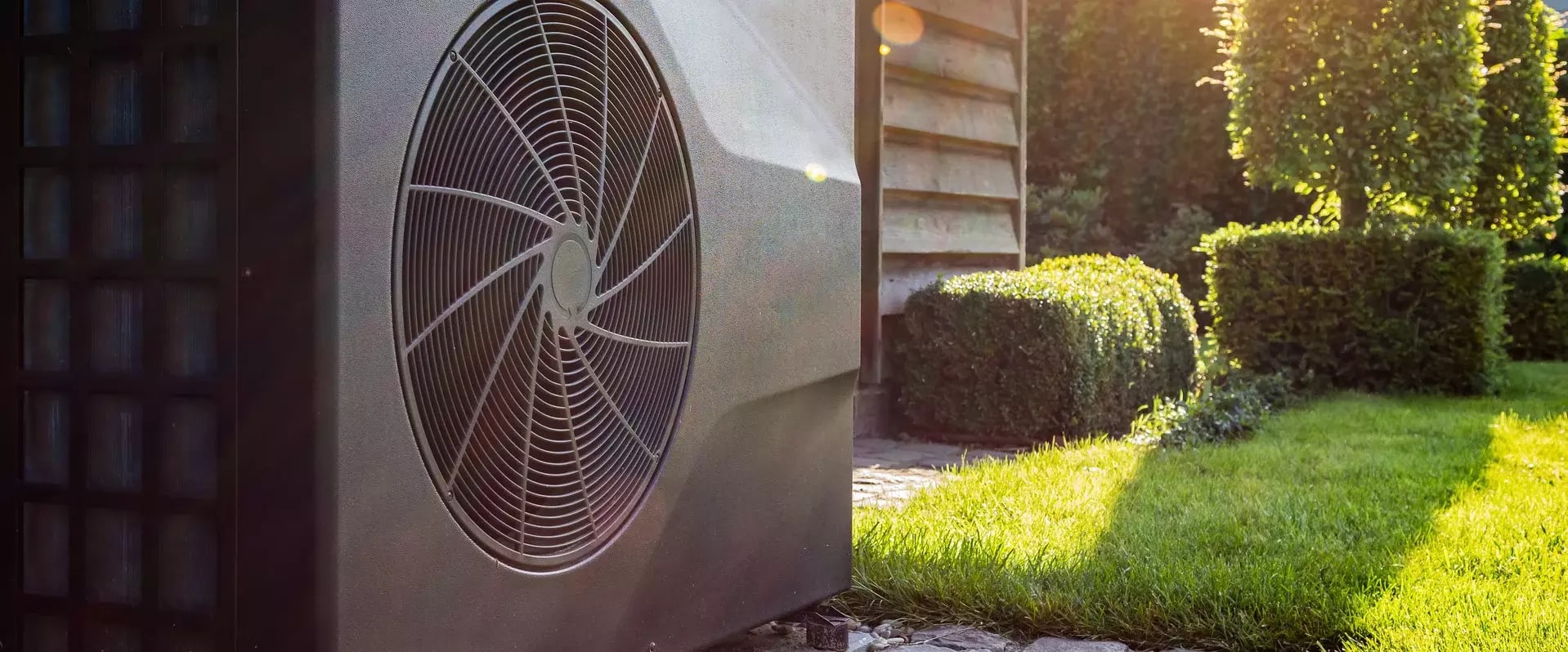 Außeneinheit einer modernen Wärmepumpe neben einem Wohnhaus, umgeben von gepflegtem Garten bei warmem Sonnenlicht.