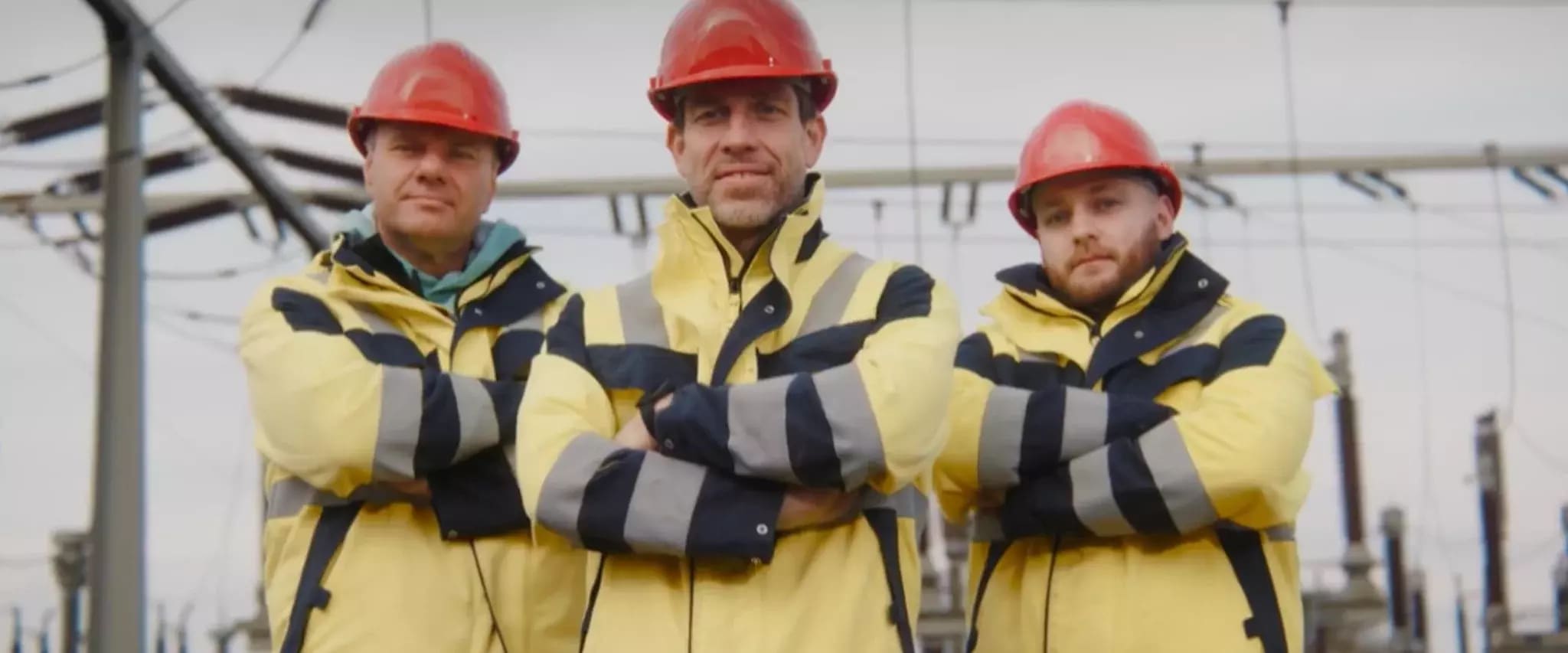 Drei Bauarbeiter in gelb-schwarzer Warnkleidung und roten Helmen stehen mit verschränkten Armen vor einer Stromanlage.