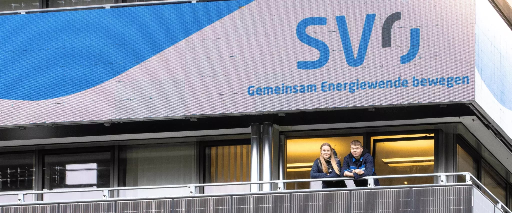 Zwei junge Menschen stehen auf einem Balkon unter einem großen LED-Schild mit dem SVO-Logo und dem Slogan 