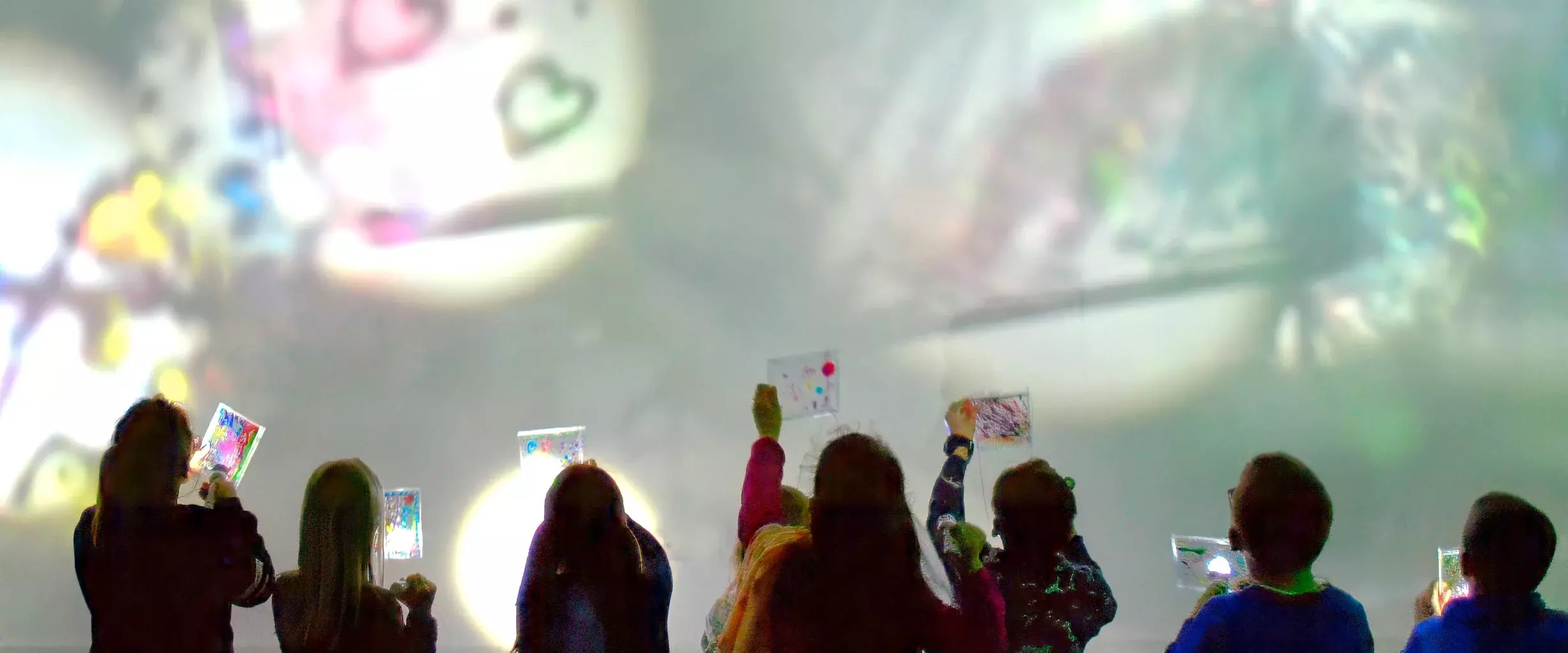 Kinder zeigen ihre bemalten Bilder vor einer großen, bunten Lichtprojektion an der Wand.
