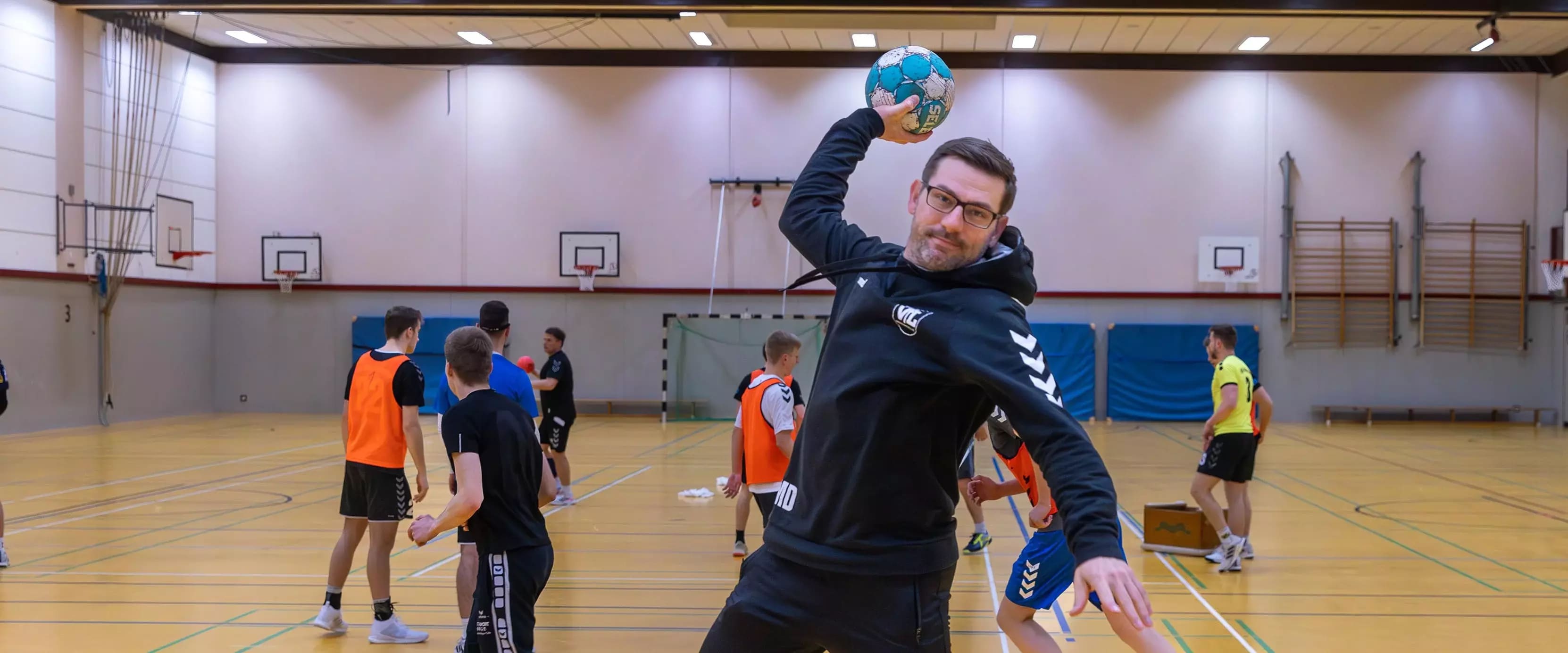 Ein Mann mit Handball in Wurfposition posiert in einer Sporthalle vor einer Gruppe junger Spieler beim Training.