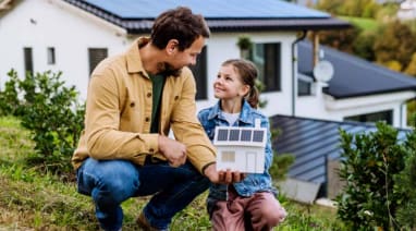 Ein Mann und ein Mädchen knien auf einer Wiese vor einem Haus mit Solarpanels und halten gemeinsam ein Modellhaus mit Solaranlage in den Händen.